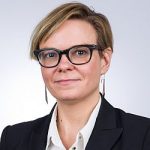 Inga Karten Portrait einer Frau mit kurzen blonden Haaren, Brille und schwarzem Blazer, vor einem hellen Hintergrund.
