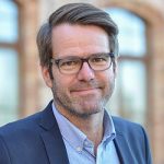 Michael Kolkmann Porträt eines Mannes mit Brille, lächelnd, in einem Anzug, vor einem unscharfen Hintergrund mit Fenstern.