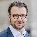 Porträt eines Mannes mit Brille, lächelnd, in Anzug, vor unscharfem Hintergrund.