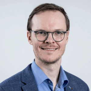 Porträt eines Mannes mit Brille, kurzen Haaren, in einem blauen Anzug und hellblauer Hemd, vor neutralem Hintergrund.