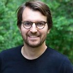 Nicolas Schwendemann Mann mit Brille und braunen Haaren lächelt vor einem grünen, unscharfen Hintergrund.