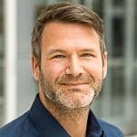 Porträt eines Mannes mit kurzen, grauen Haaren und Bart, lächelnd vor einem unscharfen Hintergrund.