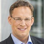 Porträt eines lächelnden Mannes mit Brille, kurzen braunen Haaren, in einem schwarzen Anzug und hellblauer Hemd.
