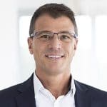 Mann mit Brille und Anzug lächelt vor einem hellen, unscharfen Hintergrund.