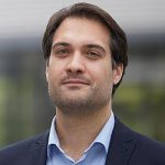 Porträt eines Mannes mit dunklen Haaren, in einem Anzug, der in die Kamera lächelt, vor unscharfem Hintergrund.