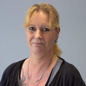 Porträt einer Frau mit blonden Haaren, die in einem schwarzen Oberteil lächelt, vor einem grauen Hintergrund.