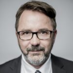 Porträt eines Mannes mit Brille, kurzen braunen Haaren und grauem Bart, in einem Anzug vor neutralem Hintergrund.