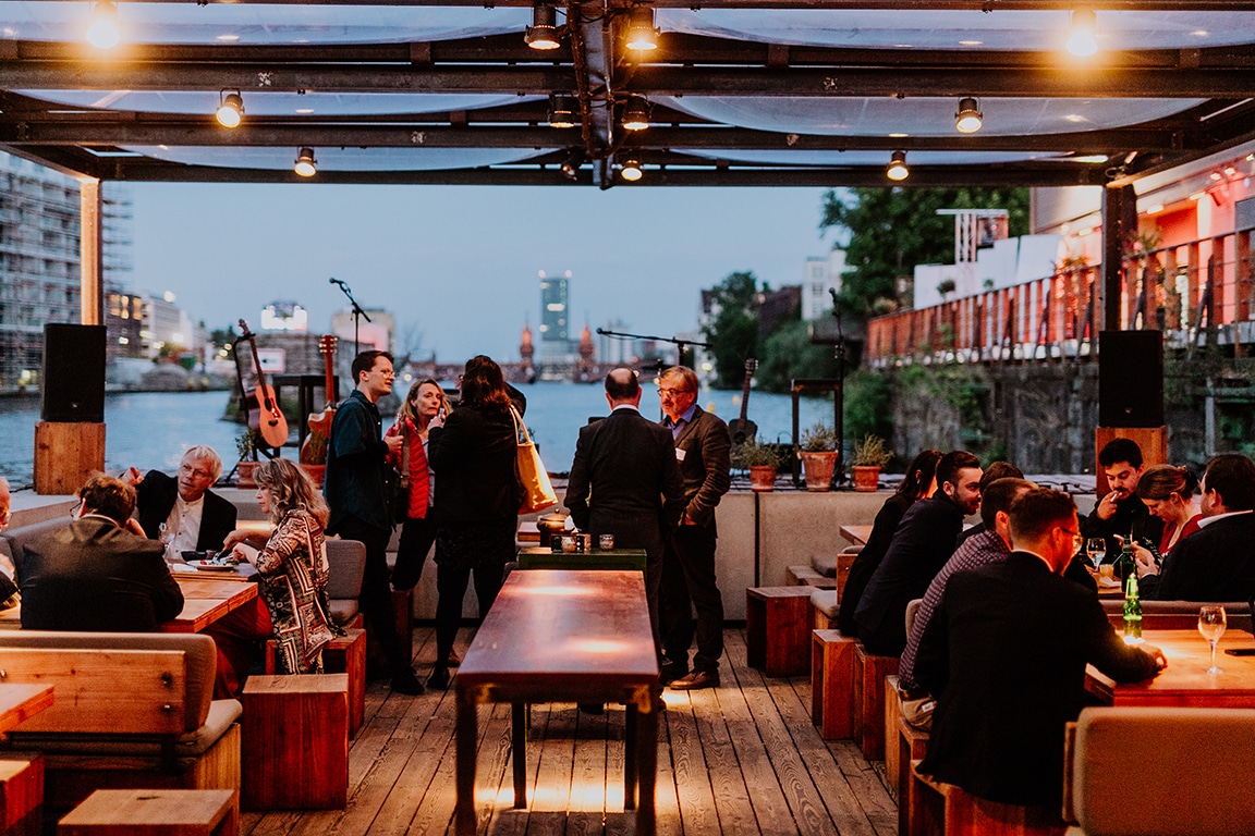 Menschen unterhalten sich in einem stilvollen Restaurant am Wasser, mit Lichtern und einer Bühne im Hintergrund.
