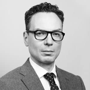 Porträt eines Mannes mit Brille, dunklen Haaren und Anzug, vor neutralem Hintergrund.