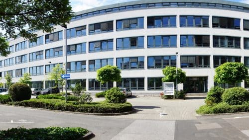 Moderne Bürogebäude mit großen Fenstern, umgeben von Bäumen und gepflegten Grünflächen.