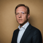 Porträt eines Mannes mit Brille, in einem Anzug, vor einem einfarbigen, braunen Hintergrund.