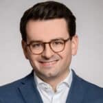 Porträt eines lächelnden Mannes mit Brille, dunklem Haar und Anzug, vor einem neutralen Hintergrund.