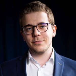 Junger Mann mit Brille, kurzem Haar und Anzug, lächelt vor einem dunklen Hintergrund.
