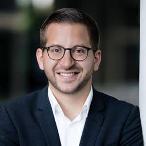 Mann mit Brille und Anzug lächelt vor unscharfem Hintergrund.