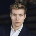 Junger Mann mit kurzem, blondem Haar, trägt einen dunklen Anzug und schaut direkt in die Kamera.