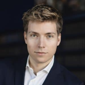 Junger Mann mit kurzem, blondem Haar, trägt einen dunklen Anzug und schaut direkt in die Kamera.