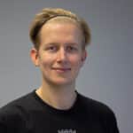 Junger Mann mit blonden Haaren und Stirnband, trägt ein schwarzes T-Shirt, lächelt vor einem grauen Hintergrund.