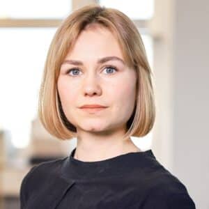 Junge Frau mit blonden, schulterlangen Haaren und blauen Augen, trägt ein schwarzes Oberteil, Hintergrund unscharf.