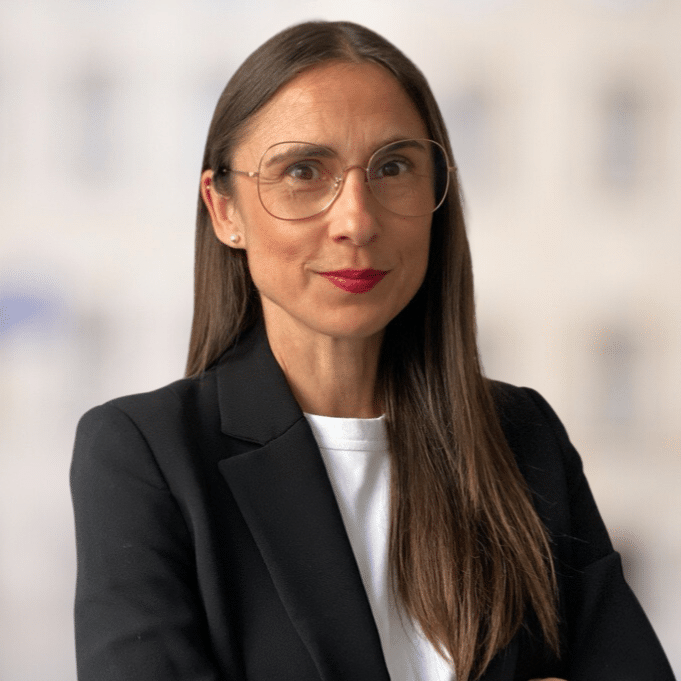Frau mit langen, glatten Haaren und Brille in schwarzem Blazer, lächelt vor unscharfem Hintergrund.