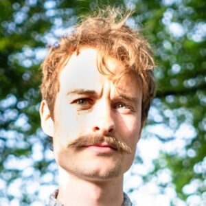Ein junger Mann mit lockigem, blondem Haar und einem markanten Schnurrbart steht vor unscharfem, grünem Hintergrund.