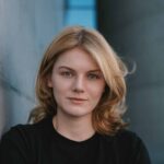 Anna Moors. Foto: Milena Radatz. Junge Frau mit blonden Haaren in schwarzem Shirt, lehnt an einer grauen Wand, mit einem modernen Gebäude im Hintergrund.