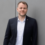 Mann in Anzug mit Hemd, steht an einer grauen Wand und schaut direkt in die Kamera.