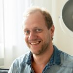 Lächelnder Mann mit blonden Haaren, trägt ein Jeanshemd, sitzt in einem hellen Raum mit Fensterlicht.