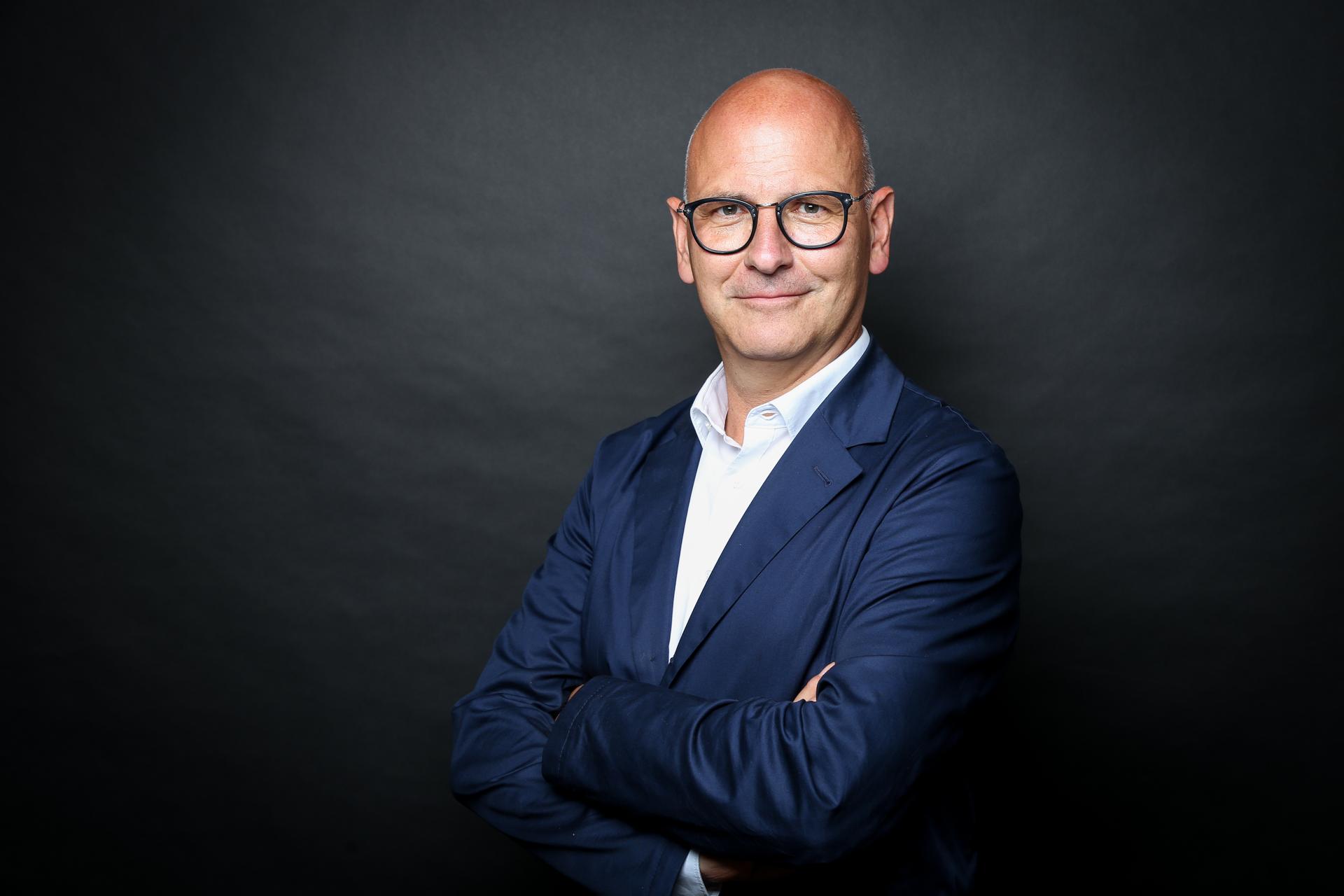 Mann mit Brille und Anzug, steht mit verschränkten Armen vor einem dunklen Hintergrund.