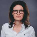 Frau mit langen, dunklen Haaren und Brille, trägt ein weißes Hemd, neutraler Hintergrund.