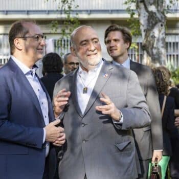 Zwei Männer im Anzug unterhalten sich in einem Garten, umgeben von weiteren Personen in formeller Kleidung.