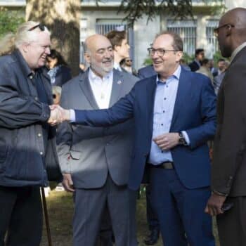 Vier Männer schütteln sich die Hände in einem Park, umgeben von Menschen und Bäumen.