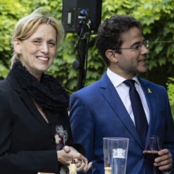 Zwei Personen in formeller Kleidung stehen lächelnd bei einer Veranstaltung im Freien, umgeben von grünen Bäumen.