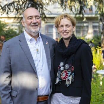 Ein Mann in einem grauen Anzug und eine Frau in schwarzer Jacke stehen lächelnd in einem Garten mit Blumen.