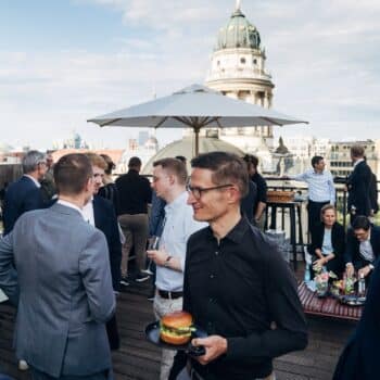 Menschen in Anzügen und legerer Kleidung stehen auf einer Dachterrasse, essen und unterhalten sich, im Hintergrund eine S...