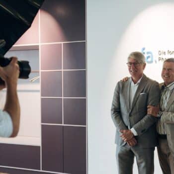 Zwei Männer posieren lächelnd für ein Foto vor einer Wand mit dem Schriftzug „Die forschenden Pharma-Unternehmen“.