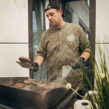 Koch mit Schürze grillt Burger-Patties auf einem Grill, umgeben von Rauch und modernen Gebäuden.