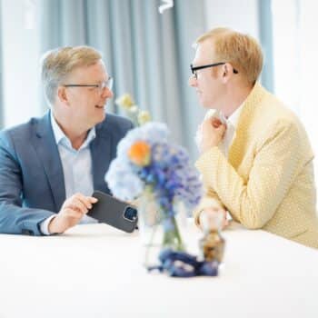 Zwei Männer in Anzügen sitzen an einem Tisch, schauen sich ein Smartphone an und unterhalten sich, mit Blumen im Vorderg...