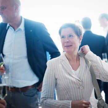 Eine Frau in gestreiftem Blazer lächelt, umgeben von Menschen in formeller Kleidung bei einer Veranstaltung.