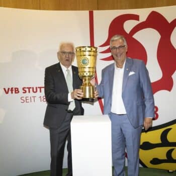 Zwei Männer halten einen großen Pokal vor einer Wand mit dem VfB Stuttgart-Logo.