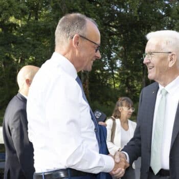 Zwei Männer schütteln sich die Hände, umgeben von Bäumen und weiteren Personen im Hintergrund.