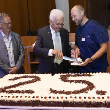 Drei Männer stehen um eine große Torte mit der Aufschrift "25." und schneiden gemeinsam ein Stück heraus.