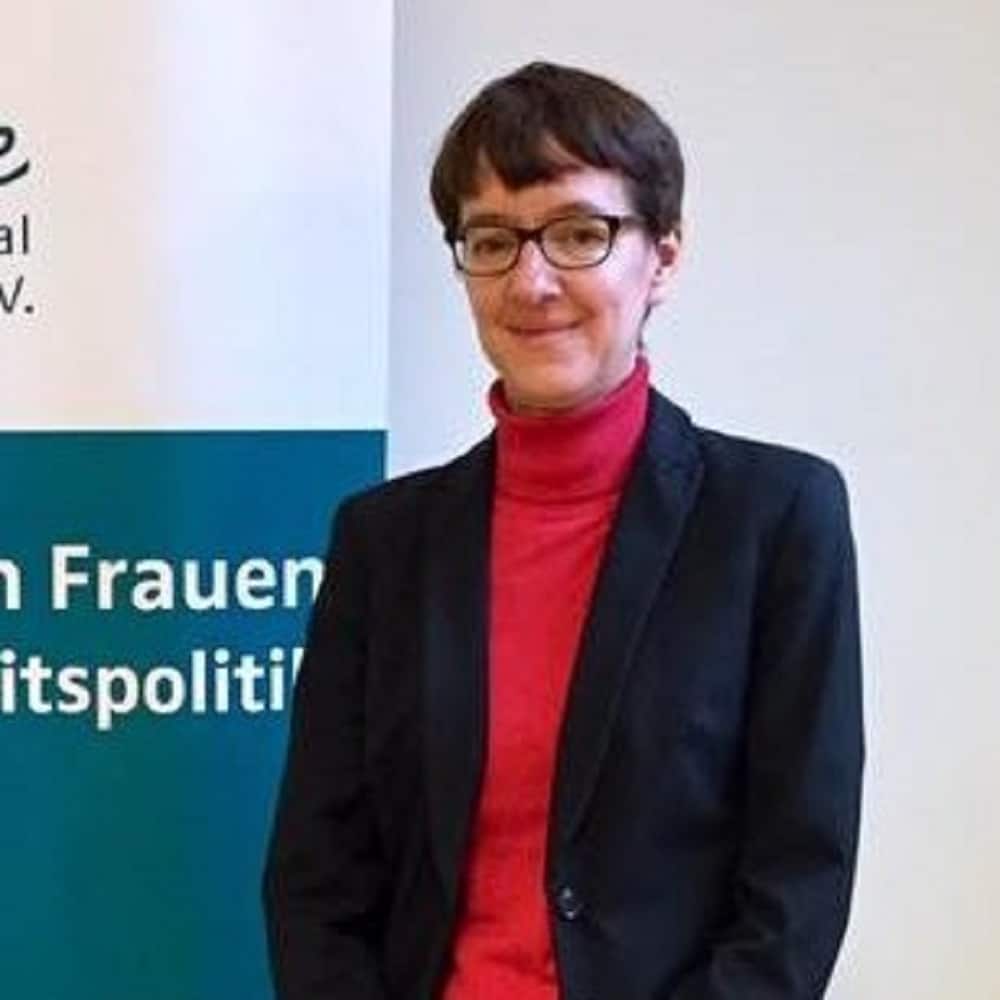 Frau mit Brille in schwarzem Blazer und rotem Pullover steht vor einem Banner mit dem Text "Frauen in der Außenpolitik".