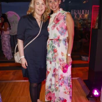 Zwei Frauen posieren lächelnd in eleganter Kleidung vor einer festlichen Kulisse mit bunten Lichtern.