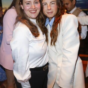 Zwei Frauen lächeln und umarmen sich, eine trägt ein weißes Oberteil, die andere einen beigen Blazer. Hintergrund mit M...