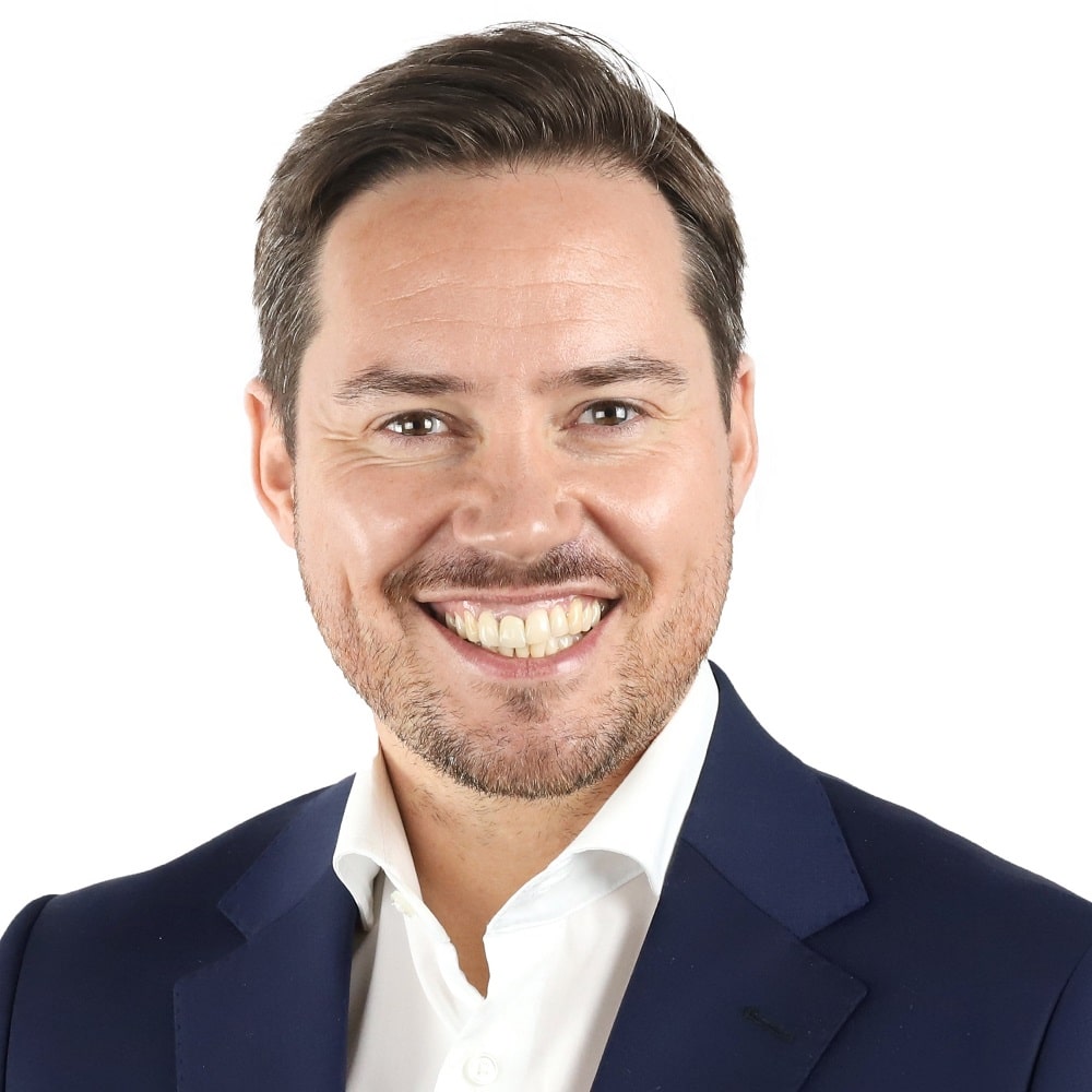 Lächelnder Mann in Anzug mit weißem Hemd vor neutralem Hintergrund.