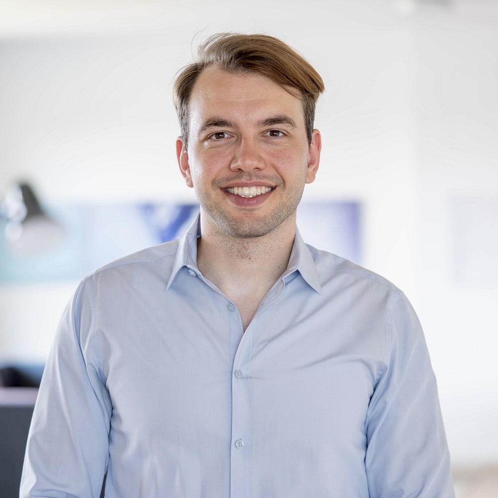 Junger Mann mit braunen Haaren und blauem Hemd, lächelt in einem modernen Büro.