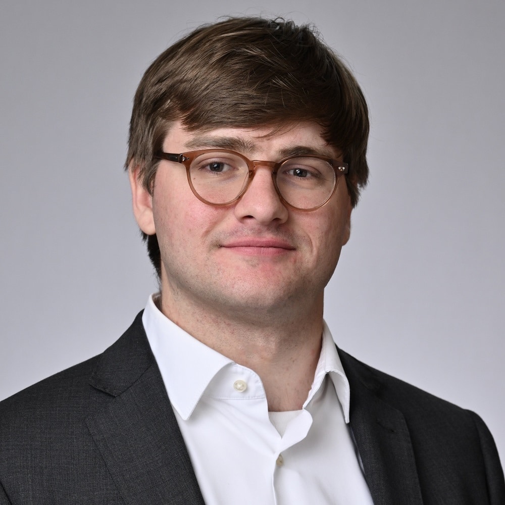 Porträt eines Mannes mit Brille, kurzen braunen Haaren, in einem Anzug mit weißem Hemd, neutralem Hintergrund.
