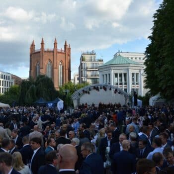 Menschenmenge bei einer Veranstaltung im Freien, umgeben von Gebäuden und Bäumen, mit einer Bühne im Hintergrund.