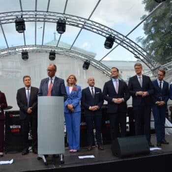 Gruppierung von neun Personen auf einer Bühne, umgeben von Musikern, unter einem transparenten Zelt bei Tageslicht.