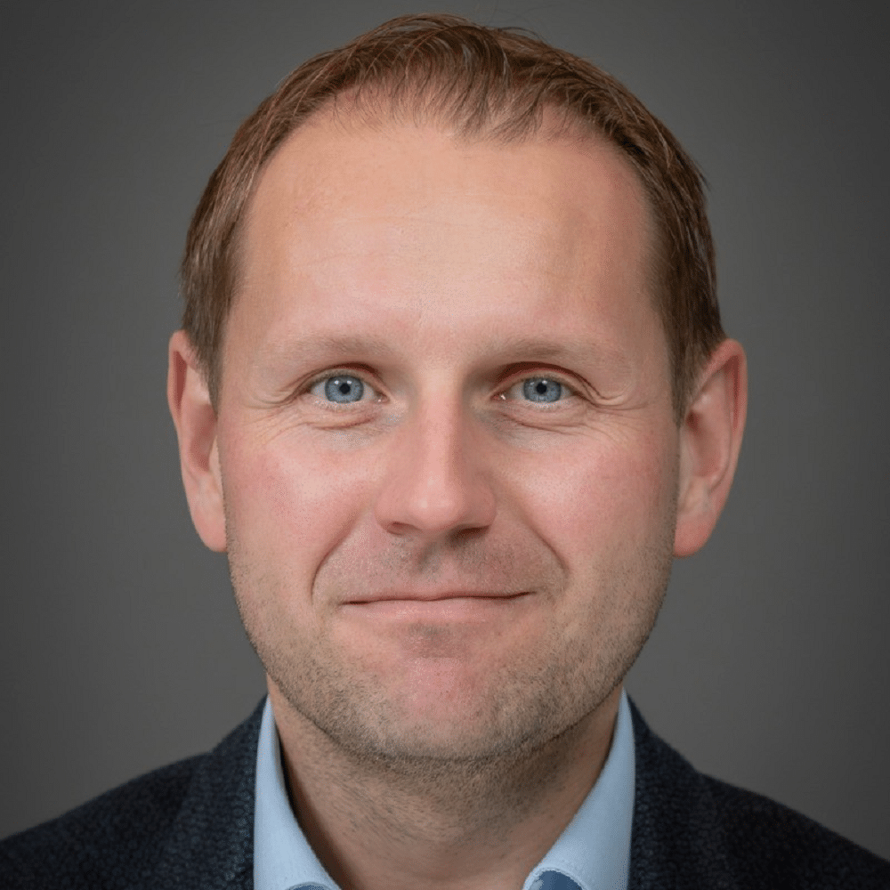 Porträt eines Mannes mit kurzen, braunen Haaren und blauen Augen, lächelnd vor einem grauen Hintergrund.